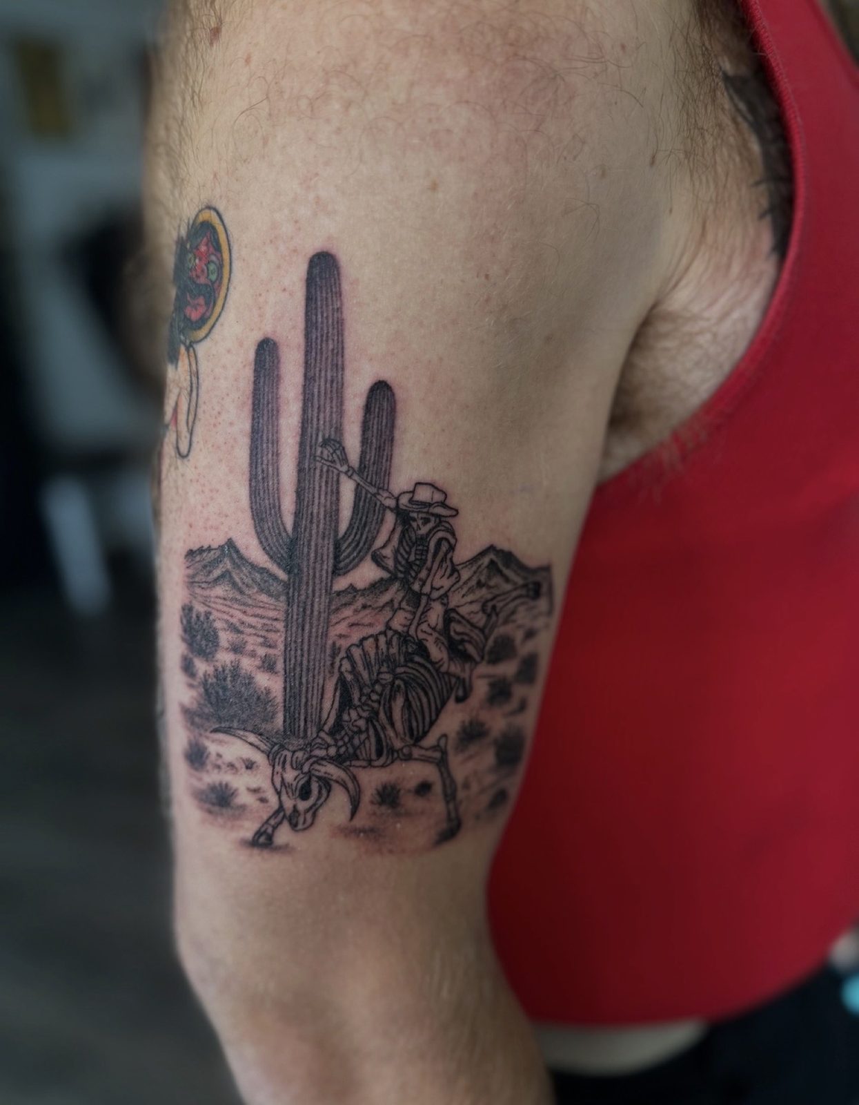 Blackwork western scene of a skeleton cowboy riding a skeleton bull beside a saguaro cactus in the desert, on the upper arm